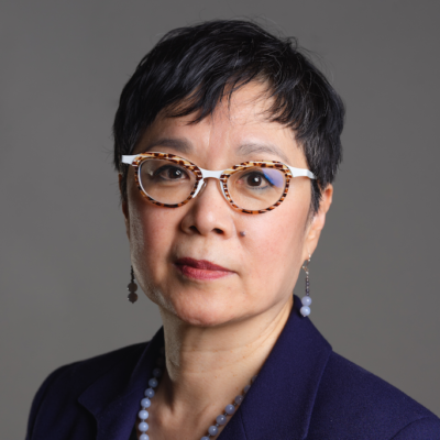 A person with short dark hair, wearing eyeglasses with a tortoiseshell frame, a purple blazer, and a beaded necklace. They are looking directly at the camera with a neutral expression, set against a plain gray background.