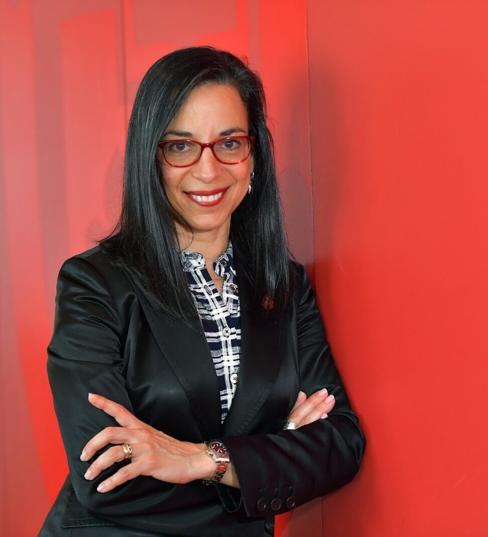 A woman with long dark hair and red glasses, wearing a black blazer over a patterned blouse, stands smiling with arms crossed against a bright red wall.