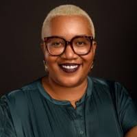 A person with short blond hair and large, round glasses smiles at the camera. They are wearing a dark green top and have dark lipstick, set against a plain dark background.