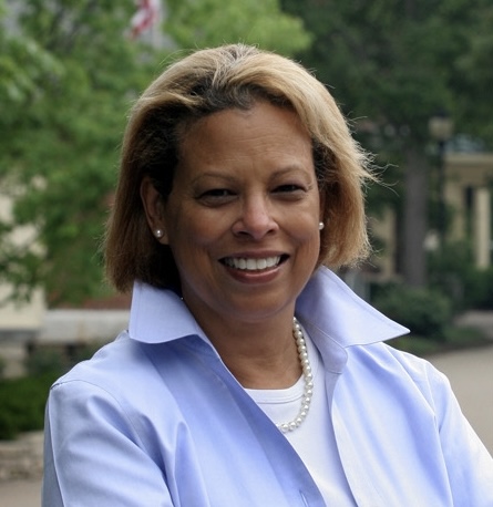 A woman with short, light brown hair smiles, wearing a light blue collared shirt, white top, and pearl necklace. She stands outdoors with greenery and blurred buildings in the background.