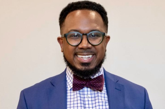 A smiling man with short curly hair, glasses, and a beard, wearing a blue suit jacket, a checkered shirt, and a maroon polka dot bow tie, stands in front of a plain light-colored background.