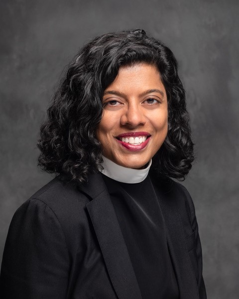 A smiling person with curly dark hair wears a black clerical jacket and a white clerical collar, posing against a gray textured background.