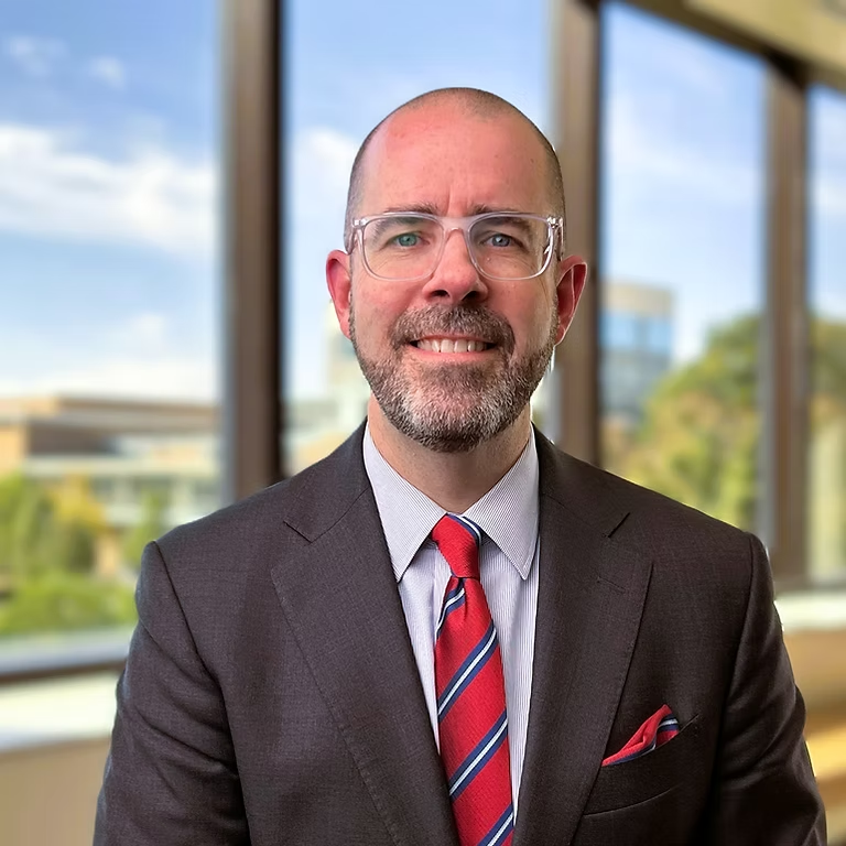 A bald man with glasses is wearing a dark suit and a red striped tie, standing indoors with large windows in the background. The setting appears to be a modern office building with a blurred outdoor view.