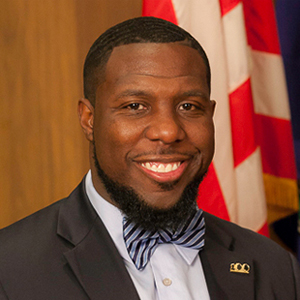 A man in a navy suit and bow tie smiles at the camera. He stands in front of a blurred U.S. flag.