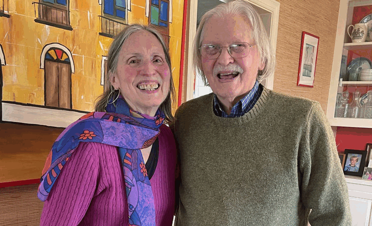 An older woman and man stand smiling together indoors. The woman wears a purple top and colorful scarf, while the man wears glasses and a green sweater. Bright artwork and a cabinet with dishes are visible in the background.