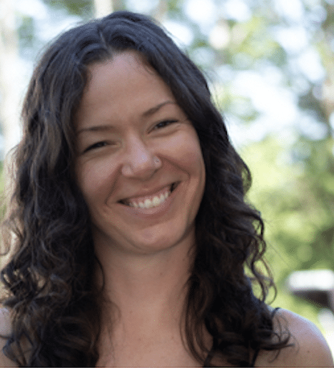 A woman with long, wavy brown hair smiles at the camera outdoors, exuding a peaceful aura inspired by Buddhism, with blurred green trees in the background.