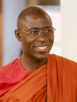 A smiling man wearing glasses and dressed in an orange robe draped over a red shirt, standing indoors.
