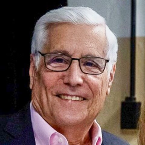 An older man with white hair and glasses is smiling. He is wearing a dark jacket over a pink shirt. The background is a plain indoor setting.