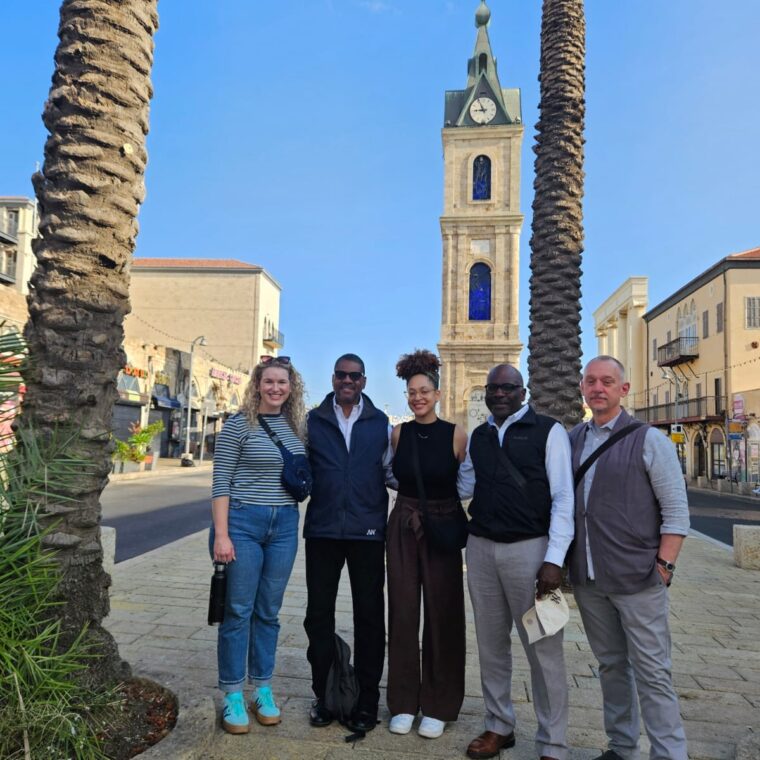 Five people stand smiling between palm trees on a sunny street, with an old clock tower in the background. They appear relaxed and happy, dressed in casual to smart-casual outfits. The sky is clear and blue.