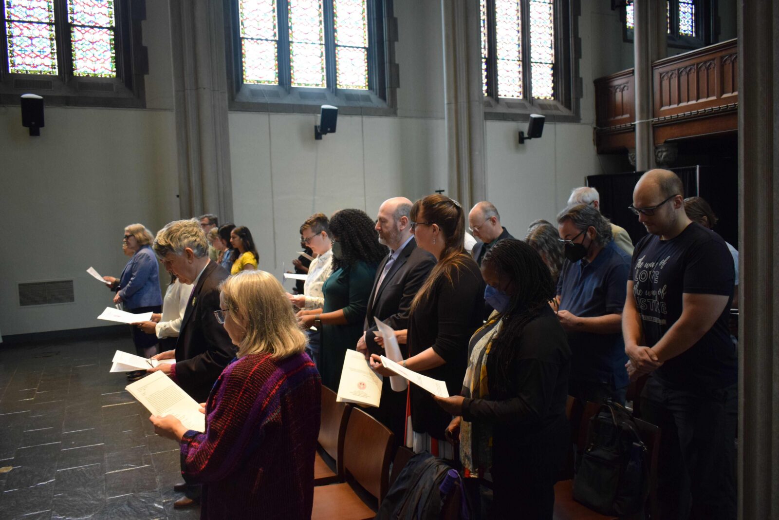 A group of people stands in rows inside a room with stained glass windows, holding papers and appearing to participate in a ceremony or event.