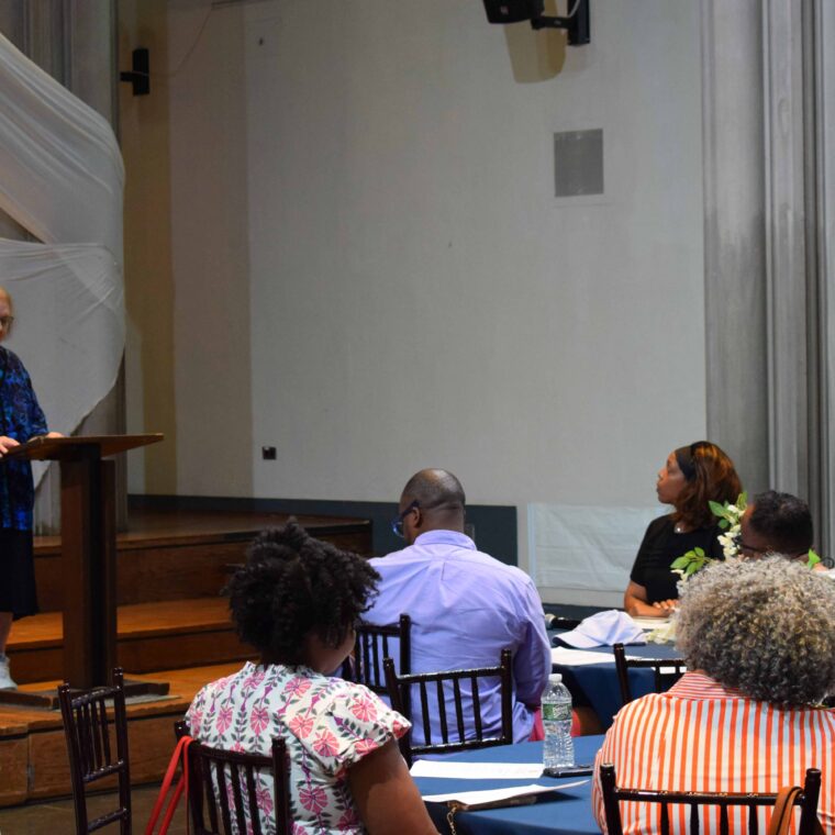 An older woman speaks at a podium on a stage while an audience sits at round tables, listening attentively in a large indoor event space.