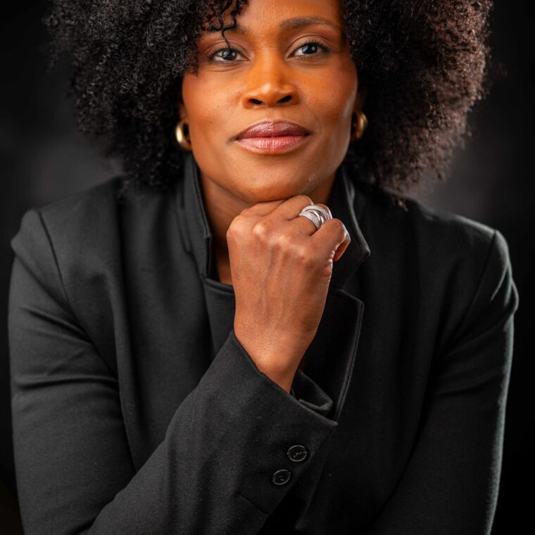 A confident woman with natural curly hair, wearing a black blazer, rests her chin on her hand and looks directly at the camera against a dark background.