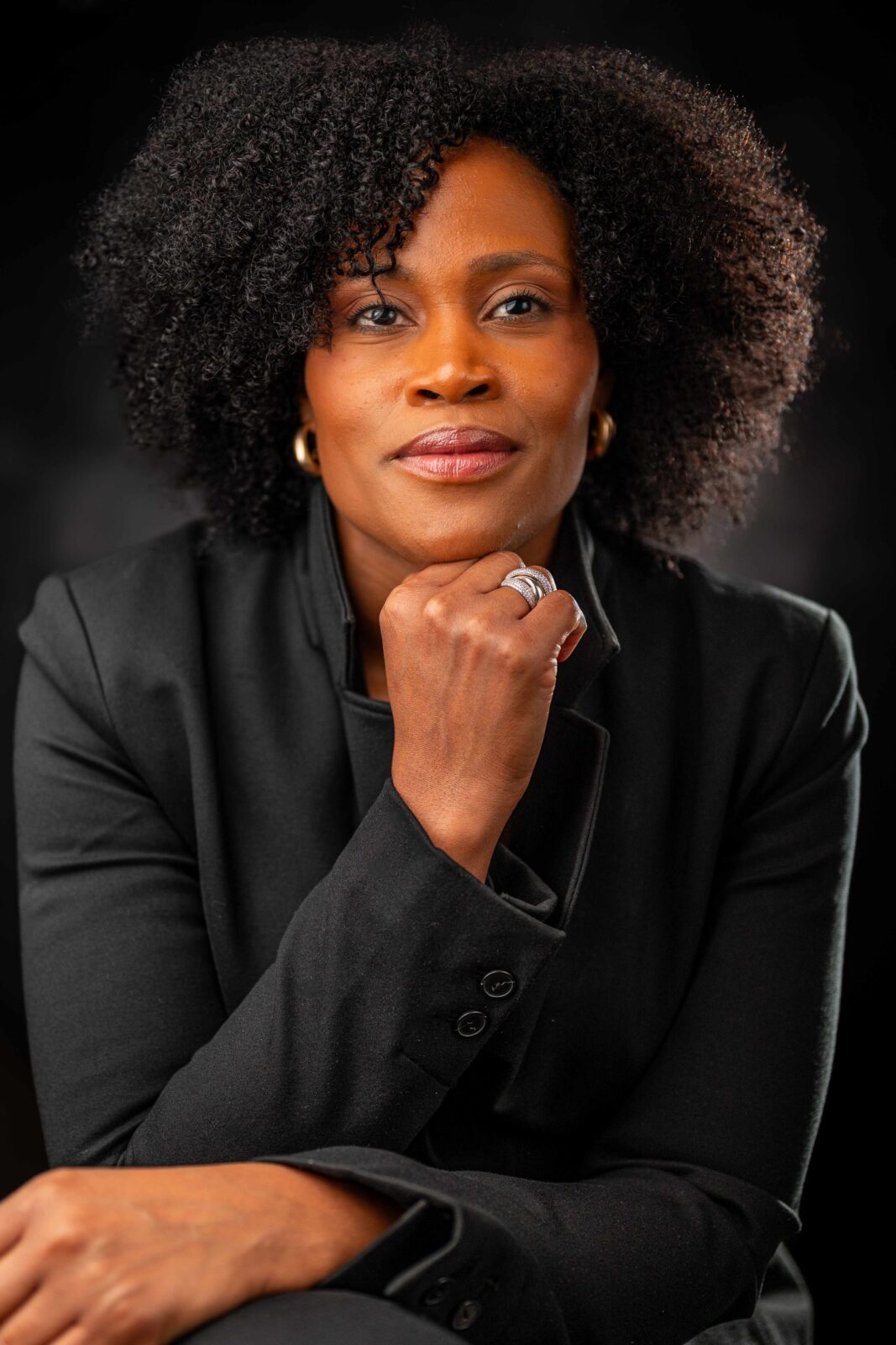A confident woman with natural curly hair, wearing a black blazer, rests her chin on her hand and looks directly at the camera against a dark background.