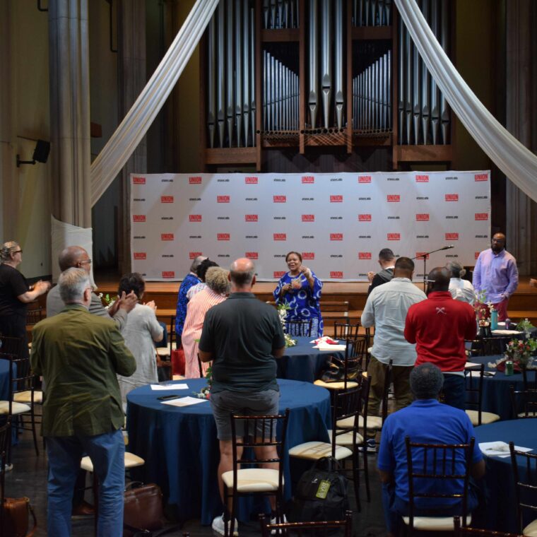 People in a hall stand and applaud a speaker at the front, where a woman addresses the audience. Round tables and chairs fill the room, with a large organ and draped fabric in the background.