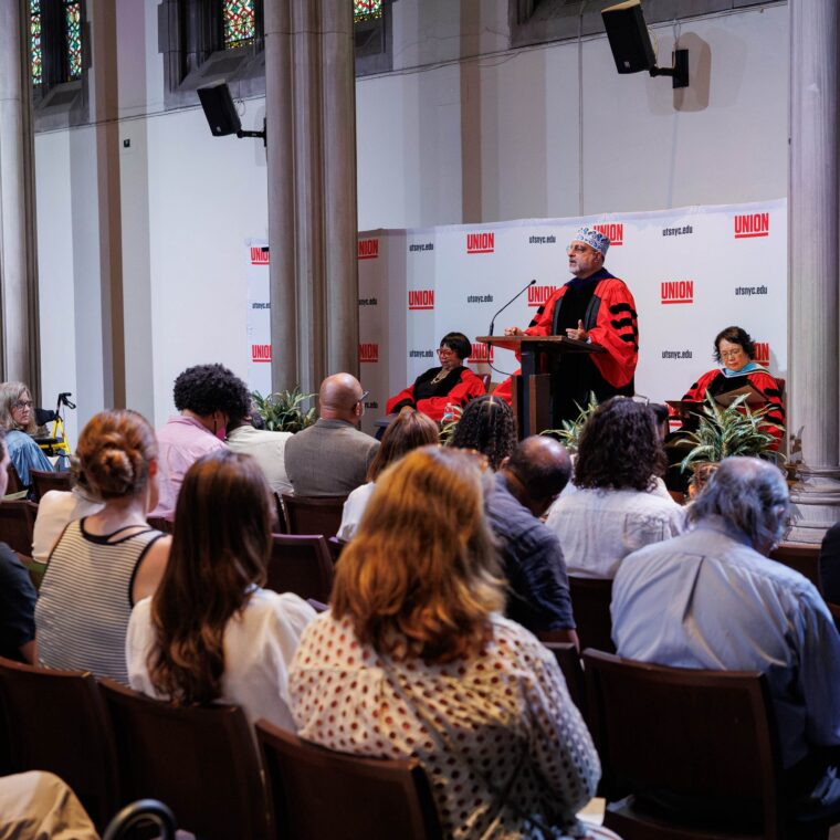 A person in academic regalia speaks at a podium during a ceremony in a hall, with an audience seated and two others in similar regalia seated onstage. A banner behind the podium reads “UNION” repeatedly.