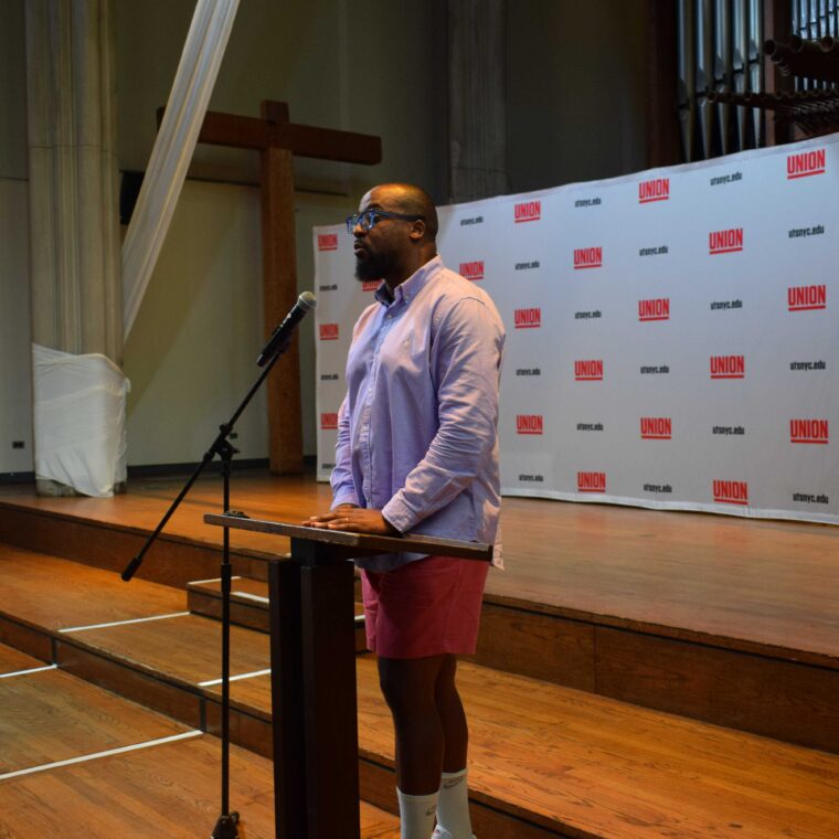 A person speaks at a podium in a large room with wooden floors, a cross, and a Union backdrop. They wear a light purple shirt, pink shorts, and glasses.