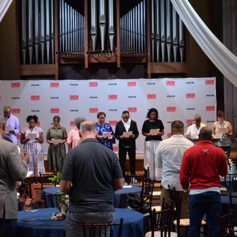 A group of people stand in a row on stage, holding papers. Audience members watch from tables. Behind them is a backdrop with “UNION” logos and a large pipe organ. Draped fabric decorates the stage area.