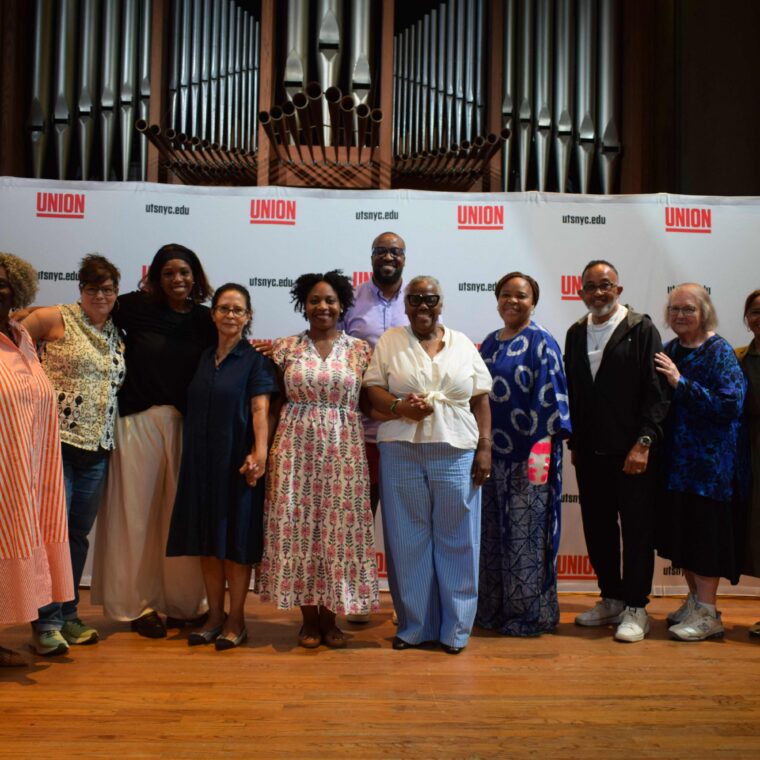 A group of twelve people stand together on a stage in front of a Union backdrop, smiling at the camera. An organ is visible in the background.