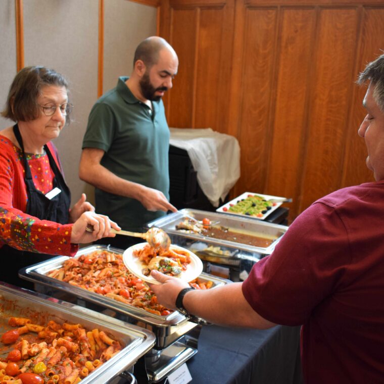 Two people serve pasta and salad from chafing dishes to a man at a buffet table in a wood-paneled room. The woman uses a spoon, while the man serves salad. The scene suggests a casual gathering or event.