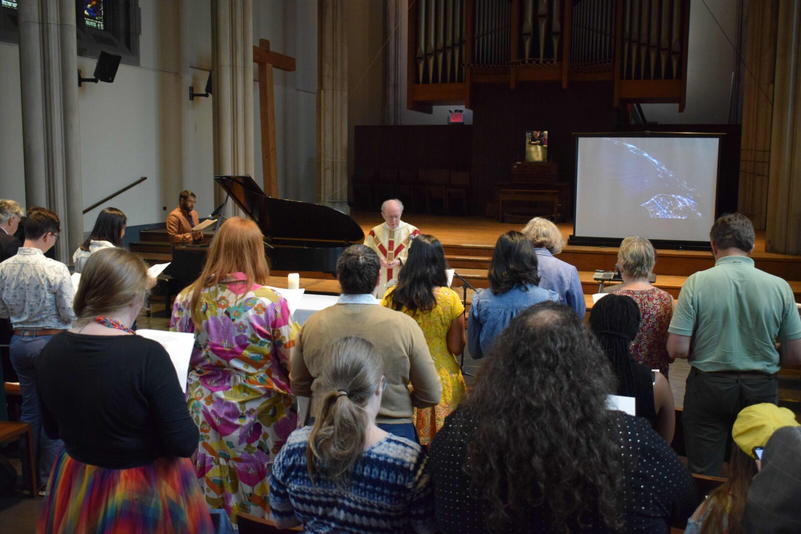 A group of people stand in a church, some holding papers. A person in robes stands at the front, with a grand piano and a large screen displaying a whale image nearby. An organ and a cross are visible in the background.
