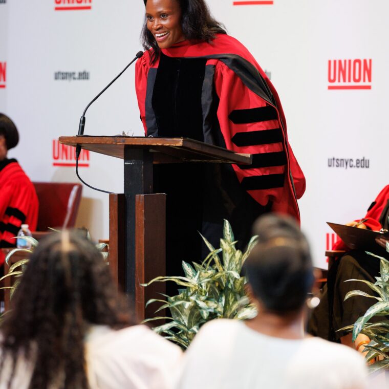 A woman in academic regalia speaks at a podium during a ceremony at Union Theological Seminary, with audience members seated and listening attentively.