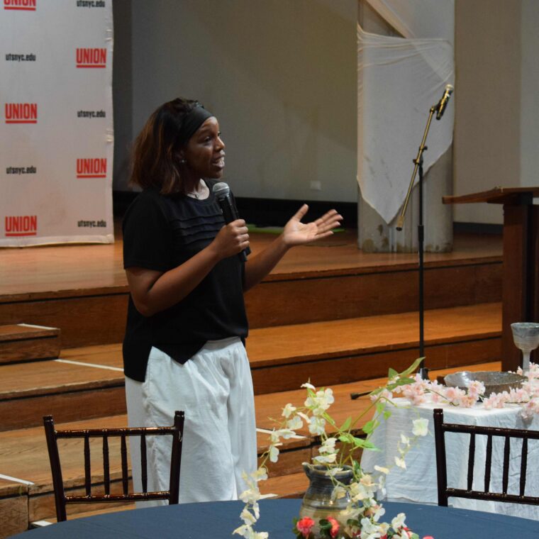 A woman stands and speaks with a microphone near a table with flowers, in front of a stage and a “Union” banner. An empty podium and mic stand are visible in the background.