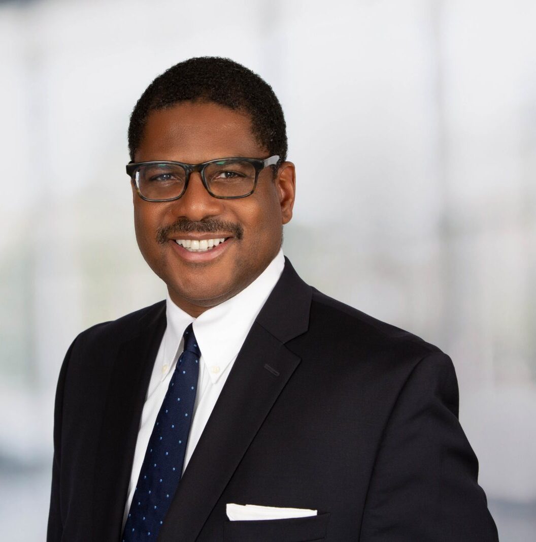 A man wearing glasses and a dark suit with a white shirt, navy tie, and pocket square is smiling in front of a blurred, bright office background.
