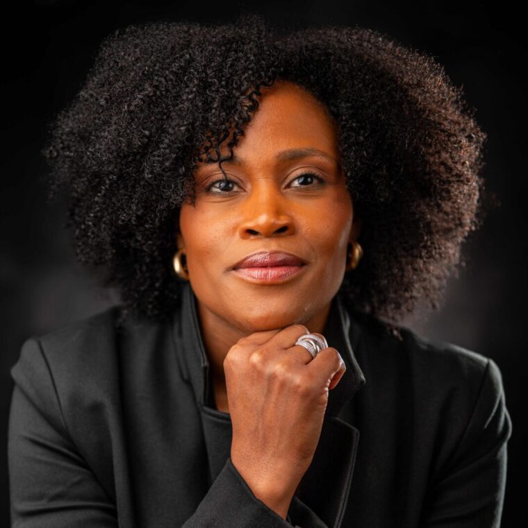 A confident woman with natural curly hair wearing a black suit sits with her hand resting on her chin, looking directly at the camera against a dark background.