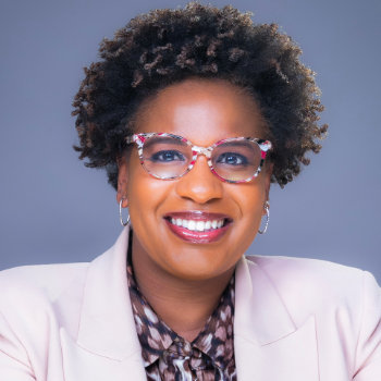 A person with curly hair smiles confidently at the camera. They're wearing colorful glasses, a light pink blazer, and a patterned blouse. The background is a gradient of gray tones.