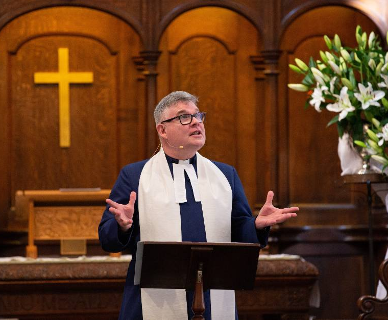 A pastor wearing glasses and a white stole speaks from a lectern in a church, with a large gold cross in the background and a bouquet of white lilies to the right.