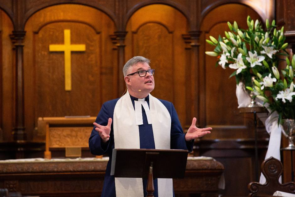 A pastor wearing glasses and a white stole speaks from a lectern in a church, with a large gold cross in the background and a bouquet of white lilies to the right.