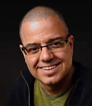 A smiling man with a shaved head, wearing glasses, a green shirt, and a dark jacket, poses against a dark background.