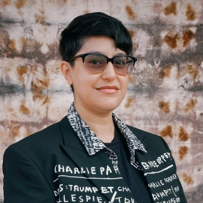 Person with short dark hair and glasses, wearing a black jacket with white text patterns. They stand against a textured, abstract brown and white background, smiling slightly at the camera.