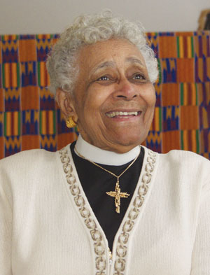 An elderly woman with gray curly hair smiles warmly. She wears a white sweater, a black shirt with a clerical collar, and a gold cross necklace. A colorful, patterned fabric hangs in the background.