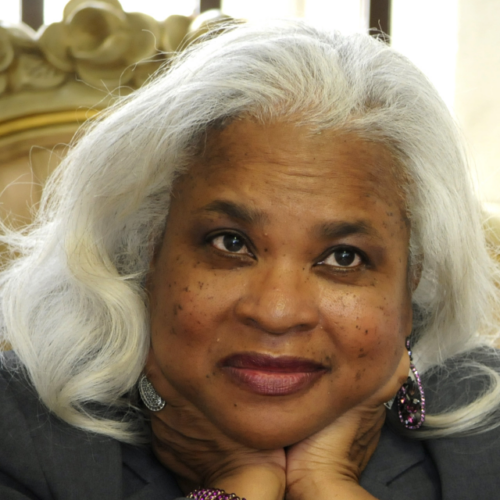 An older woman with long, wavy white hair rests her chin on her hands. She is wearing a gray blazer, large earrings, and a matching bracelet. The background is softly blurred.