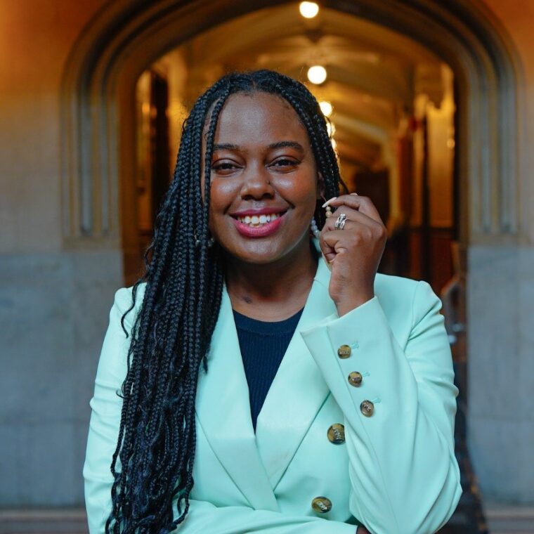 A person with long braided hair and a light green blazer smiles warmly while standing in a beautifully lit corridor with ornate architecture.