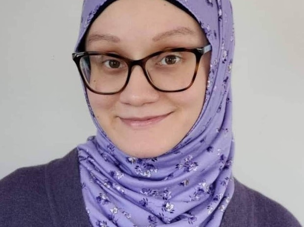 A person wearing glasses and a light purple floral hijab smiles softly at the camera. The background is plain and light-colored.
