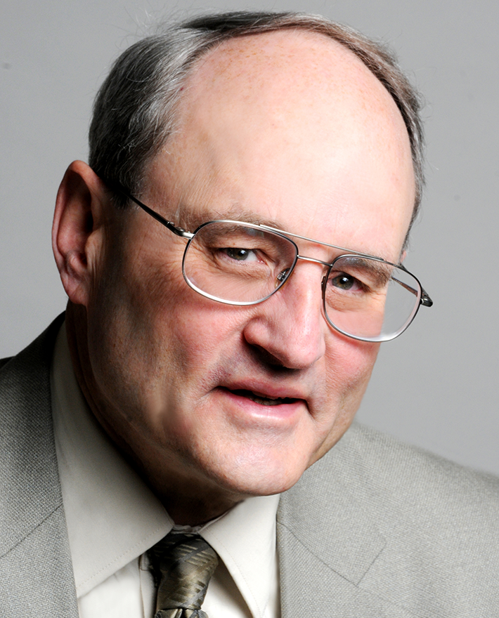 An older man with glasses and a receding hairline is wearing a light gray suit, white shirt, and patterned tie. He is facing the camera with a neutral expression against a plain background.