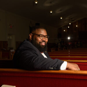 A man in a suit is smiling while sitting in a dimly lit church pew. The church interior is warmly lit with rows of wooden pews, and the walls are adorned with banners. The atmosphere is serene and contemplative.