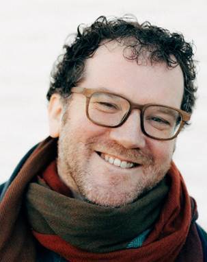 A man with curly hair and glasses smiles warmly. He is wearing a multicolored scarf and a dark jacket, standing against a light, blurred background.