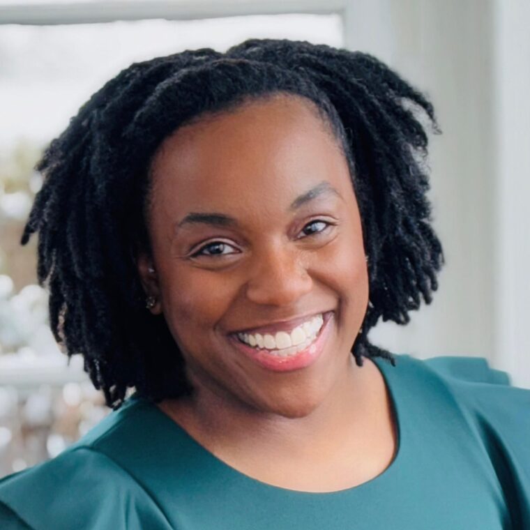 A person with a big smile and dark curly hair is wearing a green top with ruffled sleeves. They are standing in a well-lit room with a window in the background.