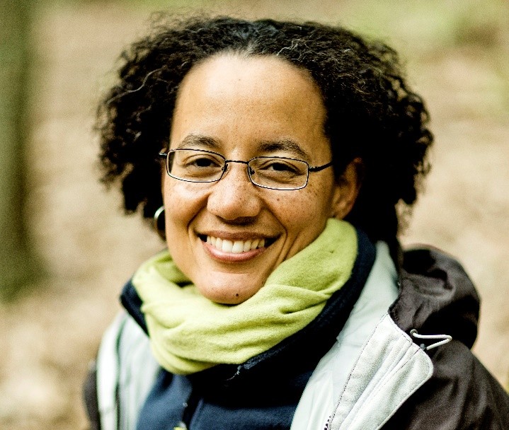 A person with curly hair and glasses smiles warmly at the camera. They are wearing a green scarf and a dark jacket. The background is blurred, hinting at an outdoor setting, possibly a forest.