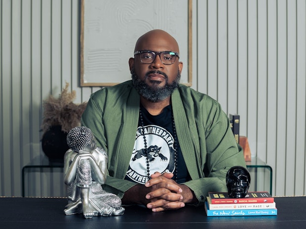 A man with glasses and a beard sits at a desk with his hands clasped. On the desk are a silver figurine, a black skull, and stacked books. He wears a green jacket over a graphic T-shirt. A textured wall art hangs behind him.