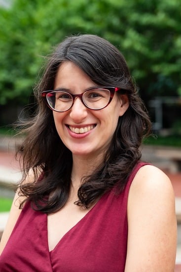 A woman with long dark hair and red glasses smiles at the camera. She is wearing a sleeveless maroon top and is outdoors, with trees and greenery in the background.