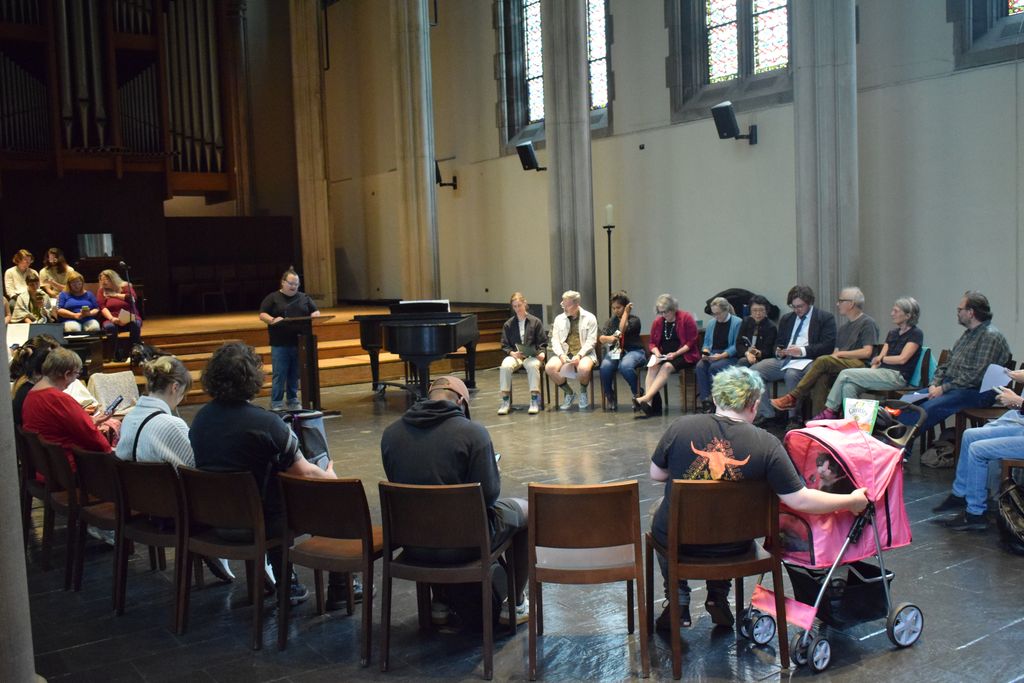 A group of people sit in a large circle in a church hall, some with papers, while a person stands and speaks. A stroller is visible in the foreground. Stained glass windows and a piano are in the background.