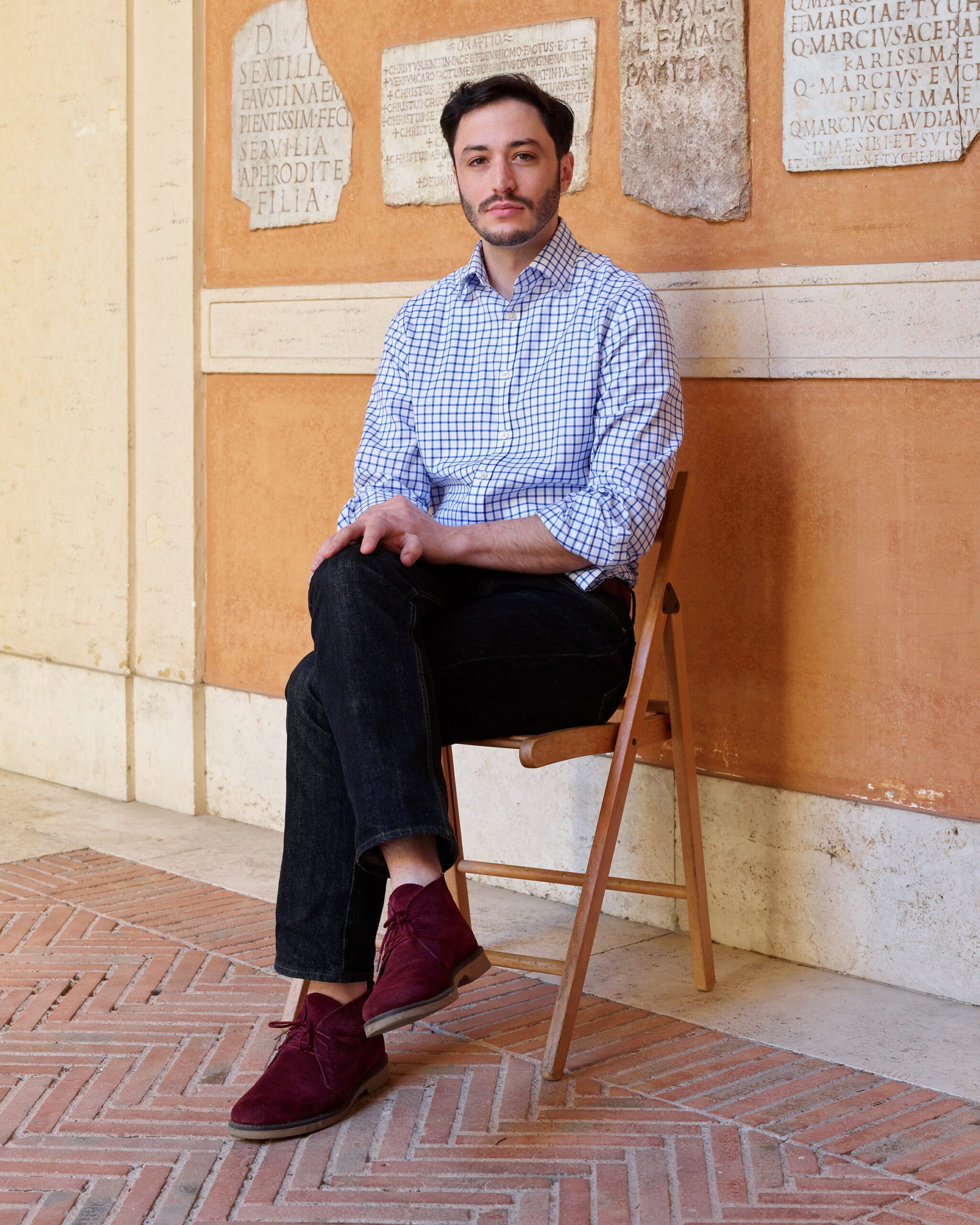 A man with short dark hair and a mustache sits on a wooden chair with his legs crossed, wearing a blue checkered shirt, dark pants, and red shoes, against a wall with ancient stone plaques and inscriptions.