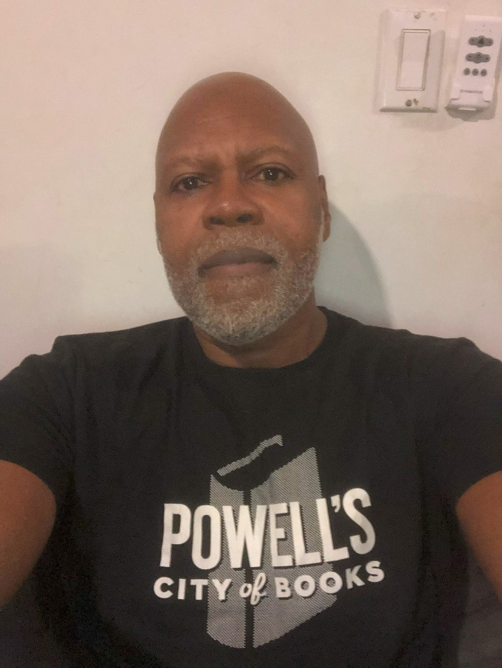 A person is wearing a black t-shirt with a white logo that reads "Powell's City of Books." They have a short beard and are sitting against a light-colored wall with a light switch and thermostat visible.