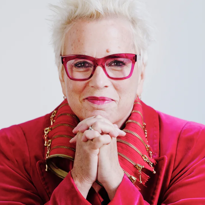 Person with short white hair, red glasses, and a bright red jacket smiles gently with hands clasped under chin against a plain light background.