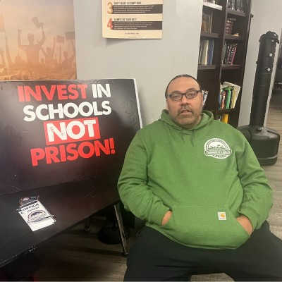 A man in a green hoodie sits next to a sign that reads, "Invest in Schools, Not Prison!" The room has bookshelves and a tall punching bag in the background. A brochure is visible on the table beside him.