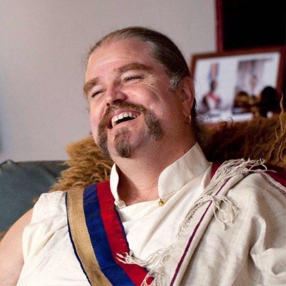 A smiling man with long hair and a mustache and beard, wearing a white robe with a colorful sash, sits on a couch. Framed photos are blurred in the background.